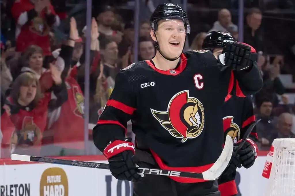 A hockey player in a black jersey with a red logo celebrates a goal, surrounded by cheering fans in a lively arena.