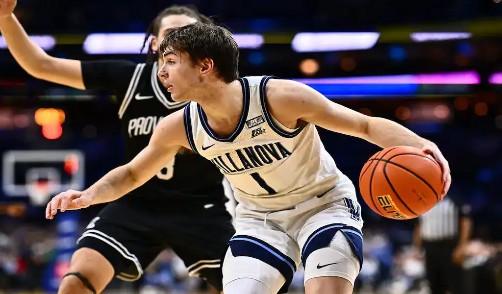 A Villanova basketball player dribbling the ball during an intense game, wearing a white and blue uniform with the number 1, while being closely guarded by an opposing player in a black Providence uniform, with a blurred crowd and court in the background.
