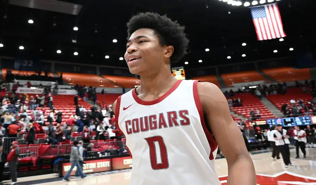 A Washington State Cougars basketball player wearing a white and red uniform with the number 0, standing on the court with a blurred crowd and American flag in the background.