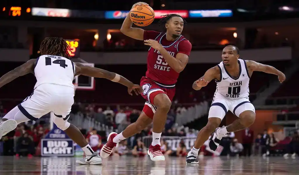 NCAAB 3 A Fresno State player in a red uniform with number 5 dribbling the ball during a game against a Utah State player in a white uniform with number 10, with a blurred crowd and court signage in the background, dated March 14, 2024.
