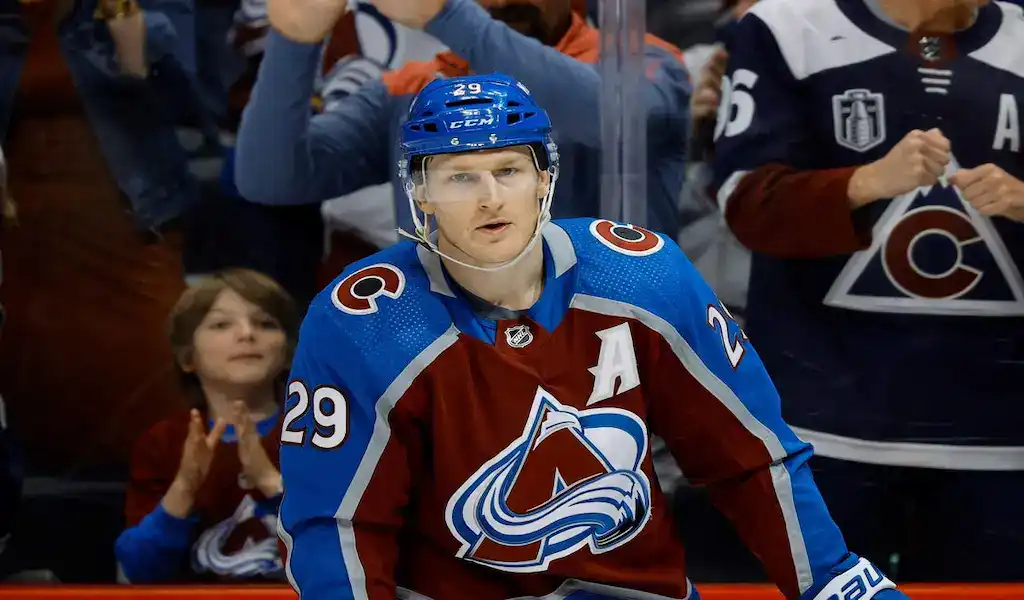 A Colorado Avalanche player in a burgundy uniform with number 29 passing the puck during a game against a Pittsburgh Penguins player in a black uniform with number 71, with a blurred crowd and rink in the background, dated March 24, 2024.