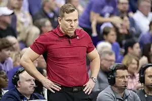 A sports coach in a red polo shirt stands with hands on hips, observing the game amidst a crowd of spectators.