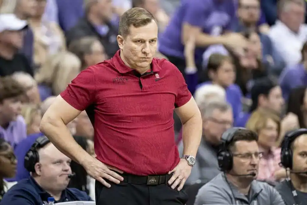 A sports coach in a red polo shirt stands with hands on hips, observing the game amidst a crowd of spectators.