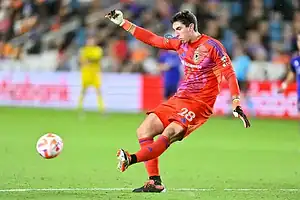 Columbus Crew goalkeeper in an orange kit kicks the ball upfield during a match.