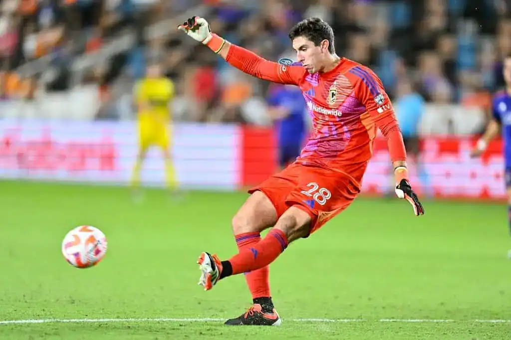 Columbus Crew goalkeeper in an orange kit kicks the ball upfield during a match.