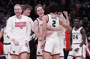 Dayton basketball players celebrate on the court after a victory, smiling and embracing.