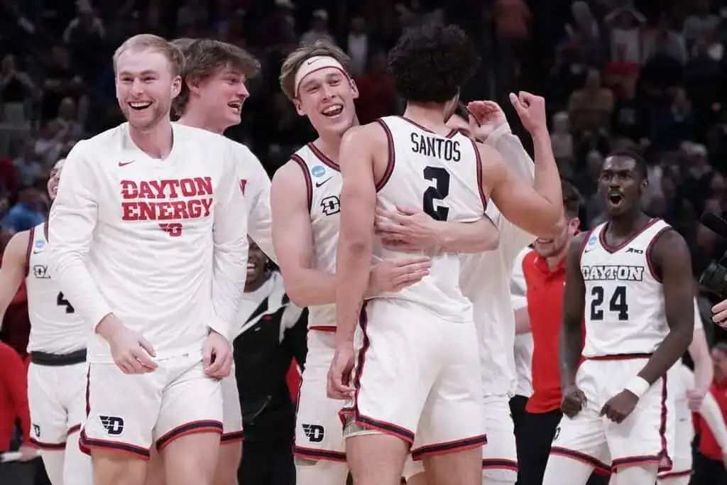 Dayton basketball players celebrate on the court after a victory, smiling and embracing.