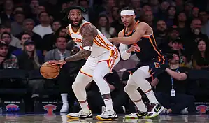 Atlanta Hawks player holds the ball under pressure from a New York Knicks defender during an NBA game.