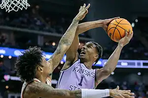 ansas State basketball player drives to the hoop under heavy defensive pressure