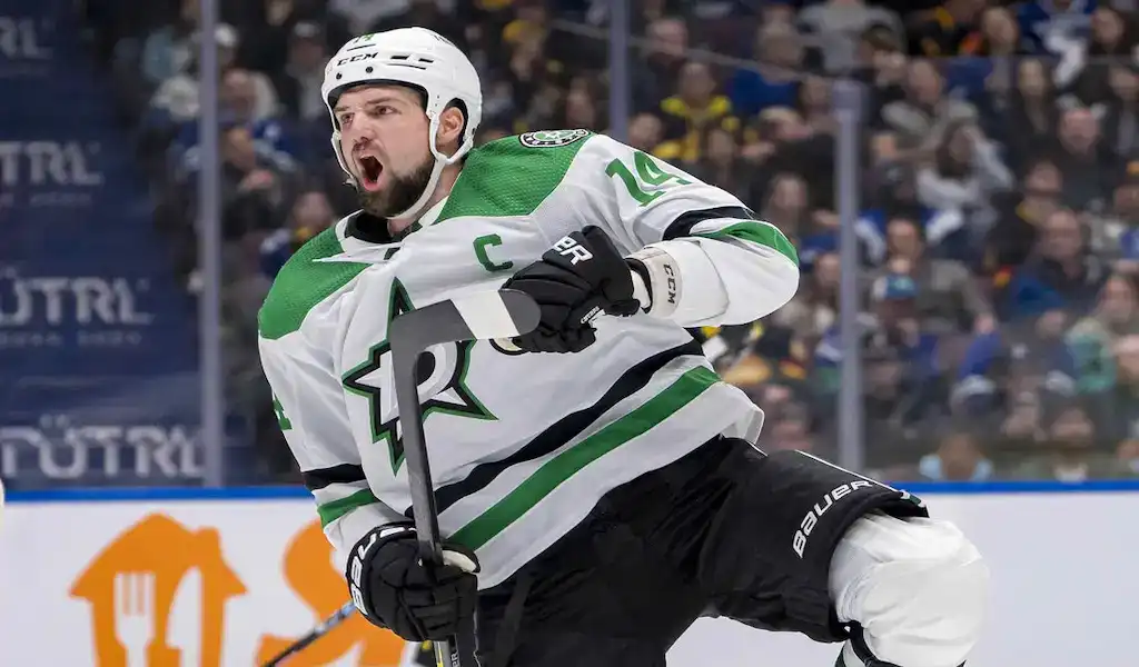 A Seattle Kraken player in a teal uniform with number 13 facing off against a Dallas Stars player in a green uniform with number 24 during a game, with a blurred crowd and ice rink in the background, dated March 30, 2024.