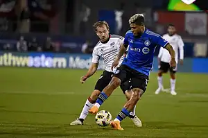 Two soccer players compete for the ball on a vibrant green field, with spectators and team banners visible in the background.