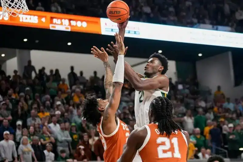 Baylor Bears player attempts a layup shot over two Texas Longhorn defenders in a packed basketball arena.