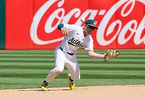 A baseball player in a white Athletics uniform fields a ball, with a vibrant Coca-Cola advertisement in the background.