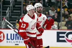 Detroit Red Wings players in white and red uniforms skate together, appearing to celebrate a play.