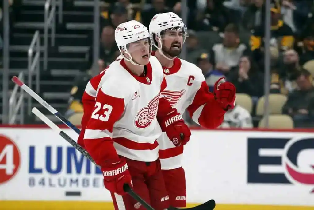 Detroit Red Wings players in white and red uniforms skate together, appearing to celebrate a play.