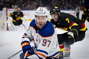 An intense hockey match scene with players in team jerseys, one maneuvering the puck while an opponent trails closely behind.