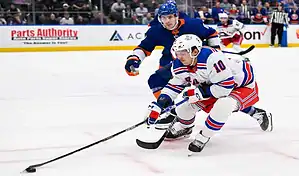 New York Rangers player controlling the puck while being defended by a New York Islanders player during an NHL game.