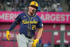 A player in a blue and yellow Milwaukee Brewers uniform runs excitedly on the baseball field, with spectators in the background.