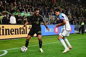 A soccer player in black kit dribbles the ball, while another player in white attempts to defend, amidst an energetic crowd.