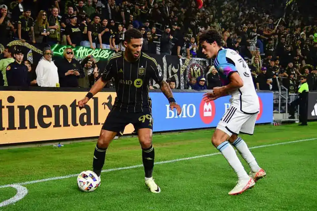 A soccer player in black kit dribbles the ball, while another player in white attempts to defend, amidst an energetic crowd.