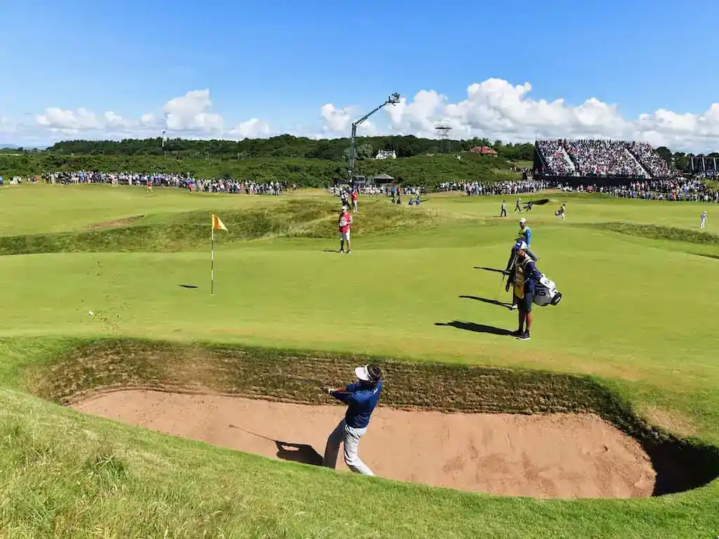 A golfer hits from a sand bunker on a sunny day at a crowded golf tournament, with spectators watching in the background.
