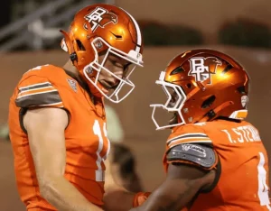 Two football players in orange uniforms celebrate on the field under stadium lights, showcasing their team's logo and gear.
