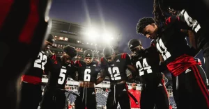 A group of football players in black and red uniforms huddle together under bright stadium lights, showing unity and focus.
