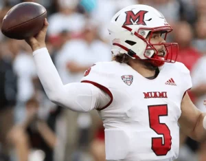 A Miami football player in a white jersey with red accents prepares to pass a football during a game.