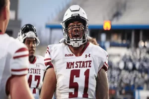 A football player wearing a white jersey with "NM STATE" and the number 51 stands confidently on the field, surrounded by teammates.