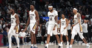 A group of basketball players in white New Orleans jerseys walk confidently on the court, surrounded by a lively crowd.