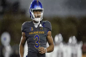 A football player in gray San Jose State uniform, wearing a blue helmet, runs on the field during a rain-soaked game night.