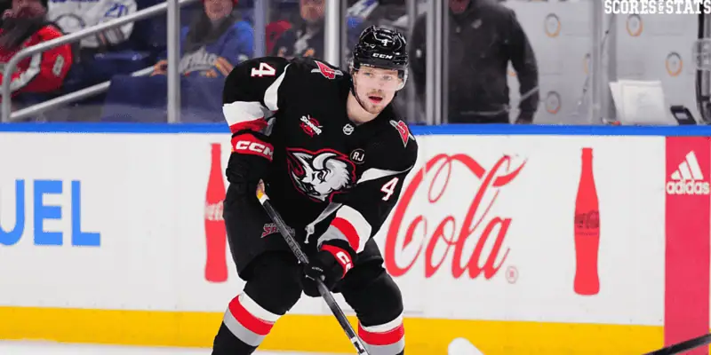  Buffalo Sabres player wearing a retro black and red jersey with a white buffalo head logo, skating with the puck during an NHL game near the boards.
