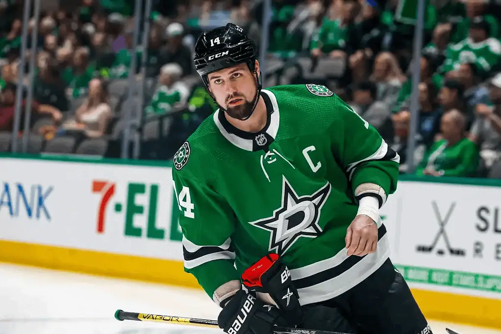 Dallas Stars vs Carolina Hurricanes Picks and Predictions January 21st 2025 2 A hockey player in a green jersey with a captain's letter skates on the ice, surrounded by fans in the stands wearing team colors.
