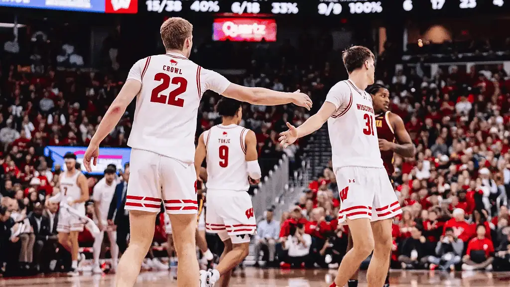 Two Wisconsin basketball players celebrate a play on the court, with fans in red jerseys cheering in the background.