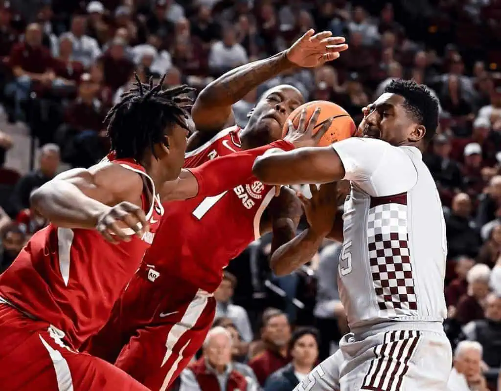 Mississippi State Bulldogs vs Texas A&m Aggies Picks and Predictions February 18th 2025 2 Two basketball players in red jerseys contest a rebound against an opponent in a white jersey, with a packed arena in the background.