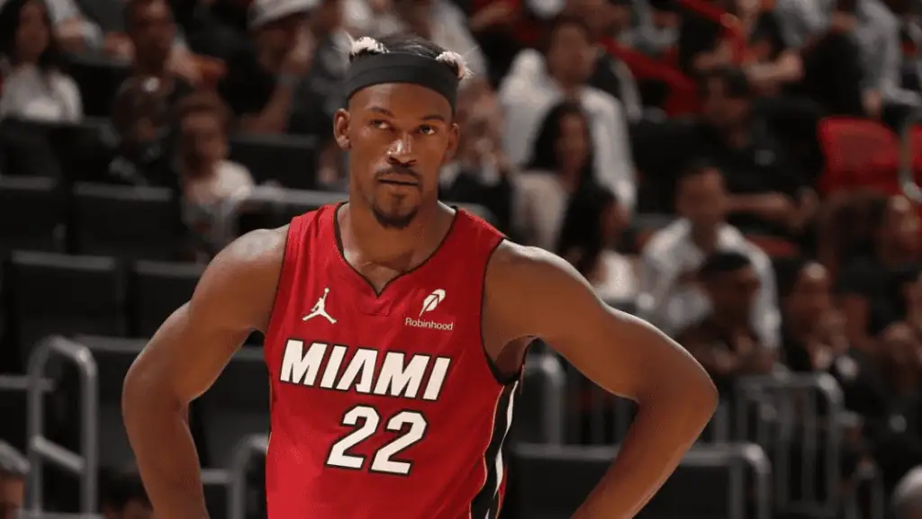 A basketball player in a red Miami jersey stands on the court, focused, with fans visible in the blurred background.