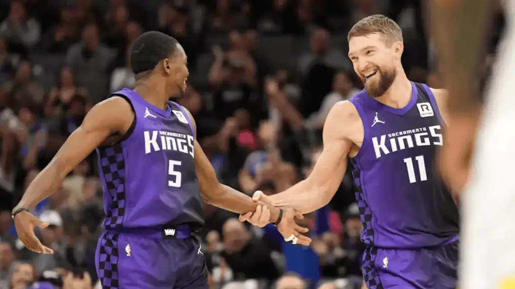 Two basketball players in purple Sacramento Kings uniforms celebrate with a high five during a game, surrounded by a cheering crowd.