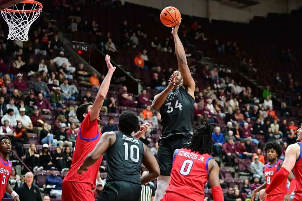 NCAAB 4 A basketball player in a black uniform leaps to shoot while two opponents in red attempt to block the shot during a game.