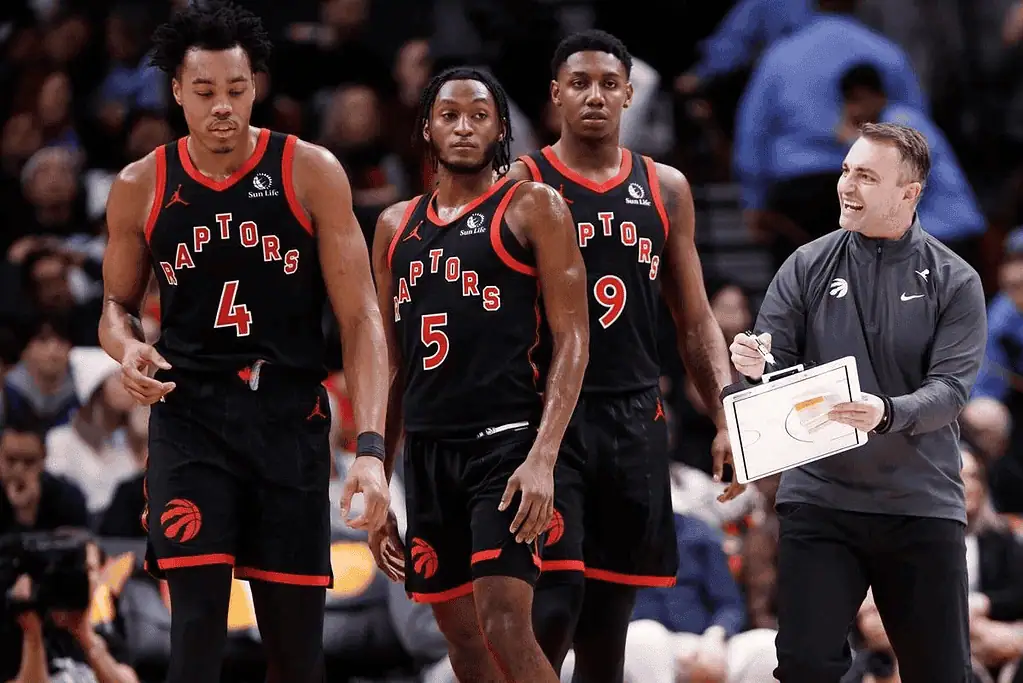 Toronto-raptors vs Boston Celtics Picks and Predictions February 25th 2025 3 A group of Toronto Raptors players in black jerseys walk alongside a coach holding a game plan on a clipboard.