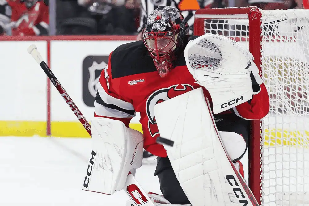 Dallas Stars vs New Jersey Devils Picks and Predictions March 4th 2025 3 A hockey goalie in a red jersey crouches at the net, holding a stick and preparing to defend against an incoming shot.