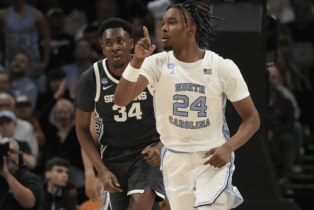 A basketball player in a white North Carolina jersey celebrates while another player in black defends, surrounded by an excited crowd.