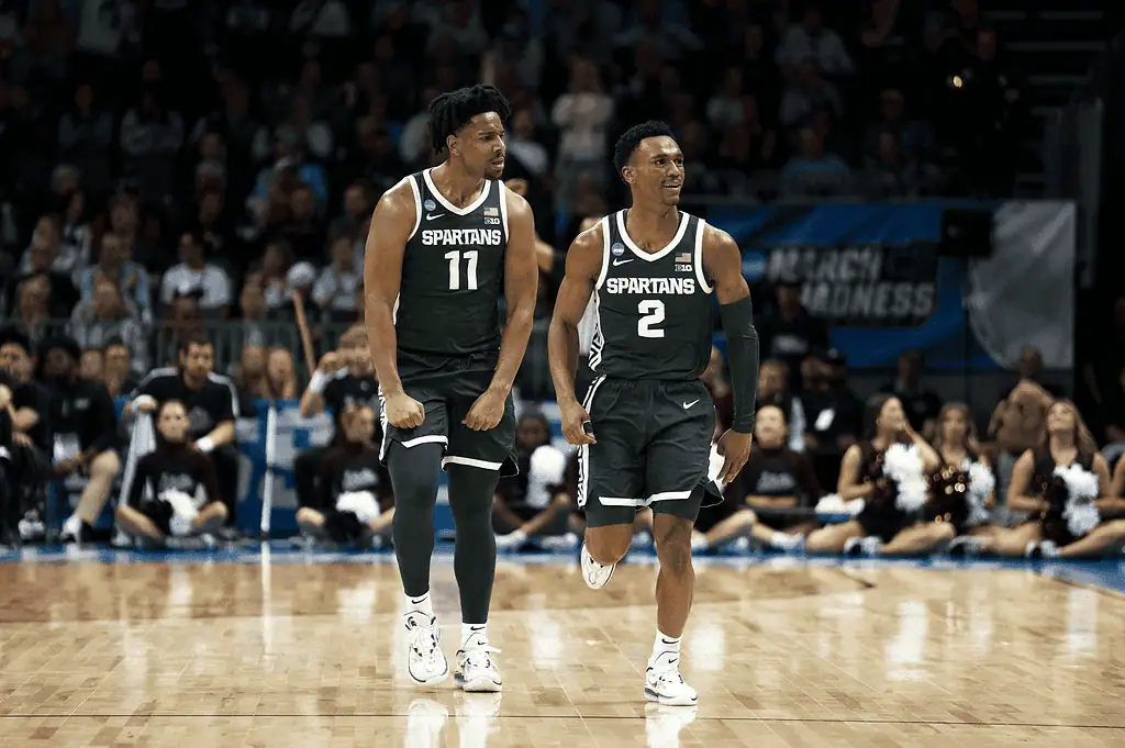 Michigan State Spartans vs Wisconsin Badgers Picks and Predictions March 2nd 2025 3 Two basketball players in green Spartan jerseys walk confidently on the court during a March Madness game, with fans in the background.