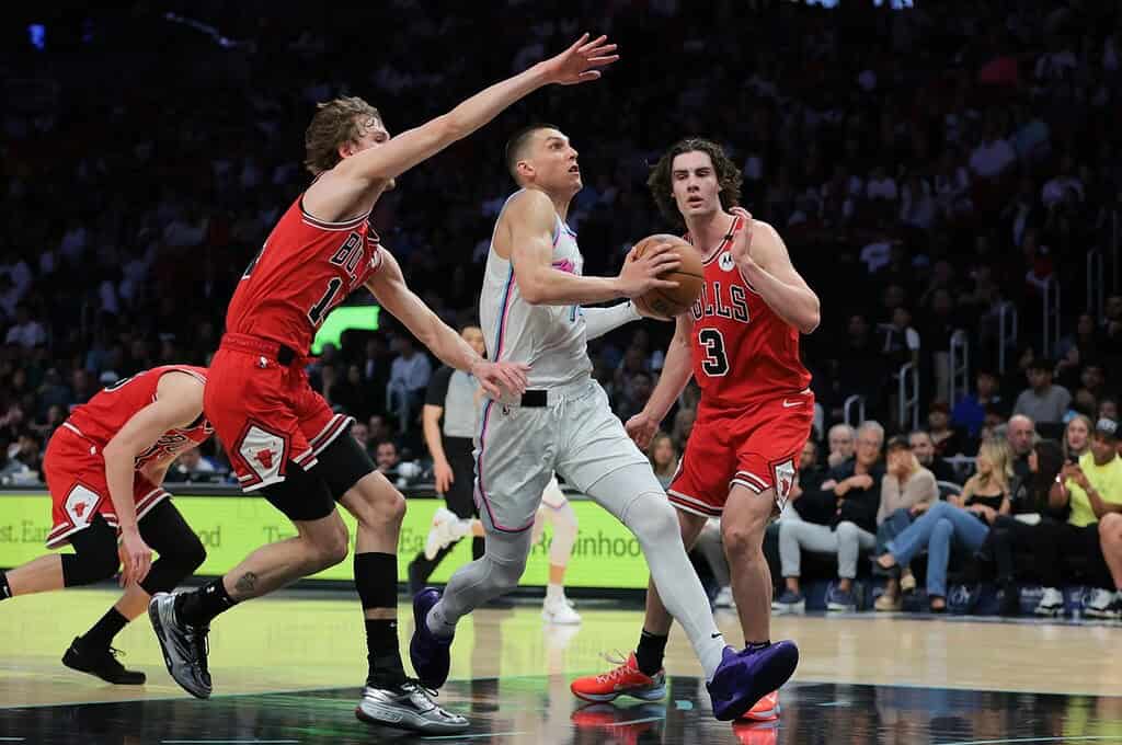 A player in a white jersey dribbles past two defenders in red jerseys during an intense basketball game, with spectators in the background.