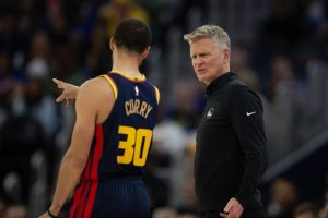 NBA player Stephen Curry, wearing a Golden State Warriors jersey, gestures while engaging in conversation with a coach during a game.