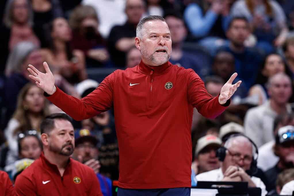 A coach in a red jacket gestures passionately during a basketball game, while fans watch intently in the background.