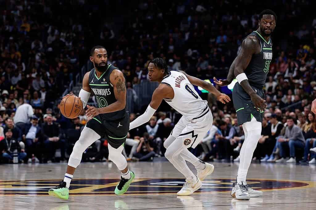 Two basketball players from opposing teams in action on the court. One dribbles the ball while the other defends. The crowd watches intently.