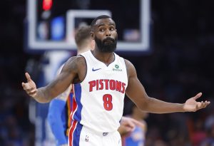 A Pistons player in a white jersey with the number 8 holds out his arms in disbelief during a game, with teammates in the background.