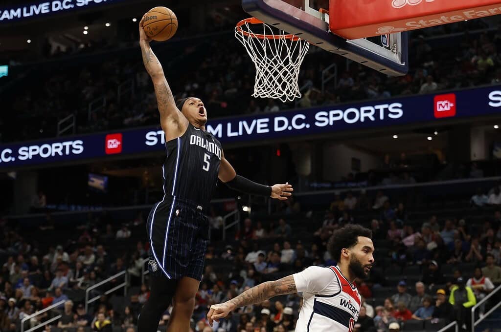 A basketball player in a blue Orlando jersey leaps for a dunk, showcasing athleticism above an opponent in a white jersey.