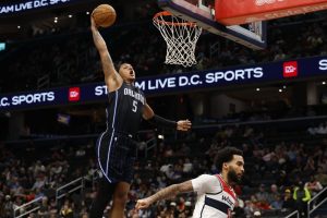 A basketball player in a blue Orlando jersey leaps for a dunk, showcasing athleticism above an opponent in a white jersey.