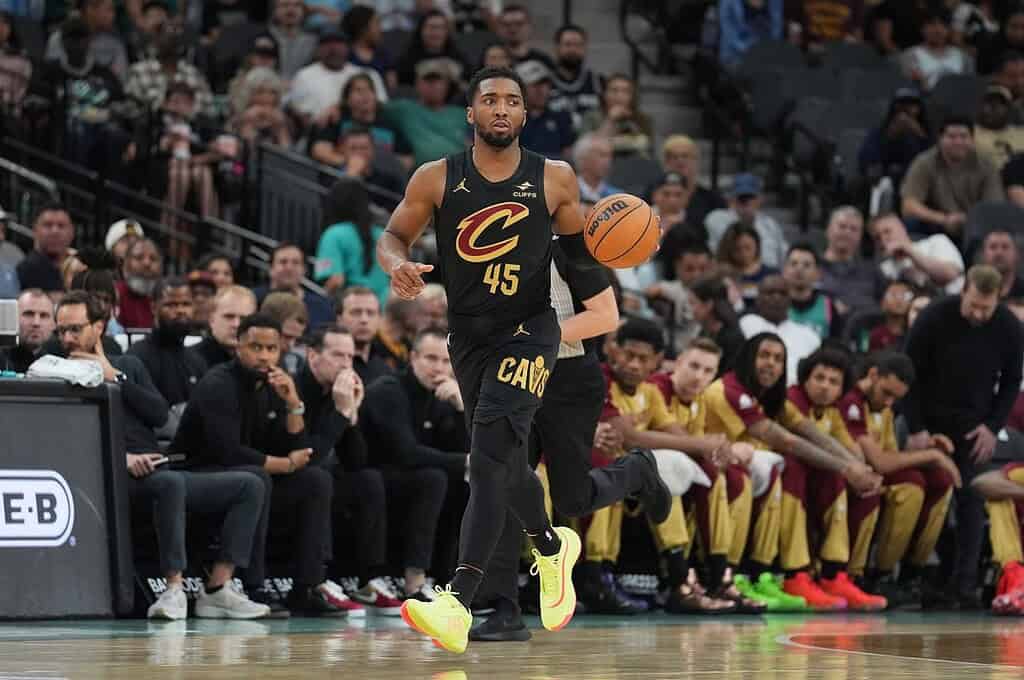 A basketball player in a black jersey with "Cavs" logo dribbles the ball during a game, with spectators and teammates in the background.